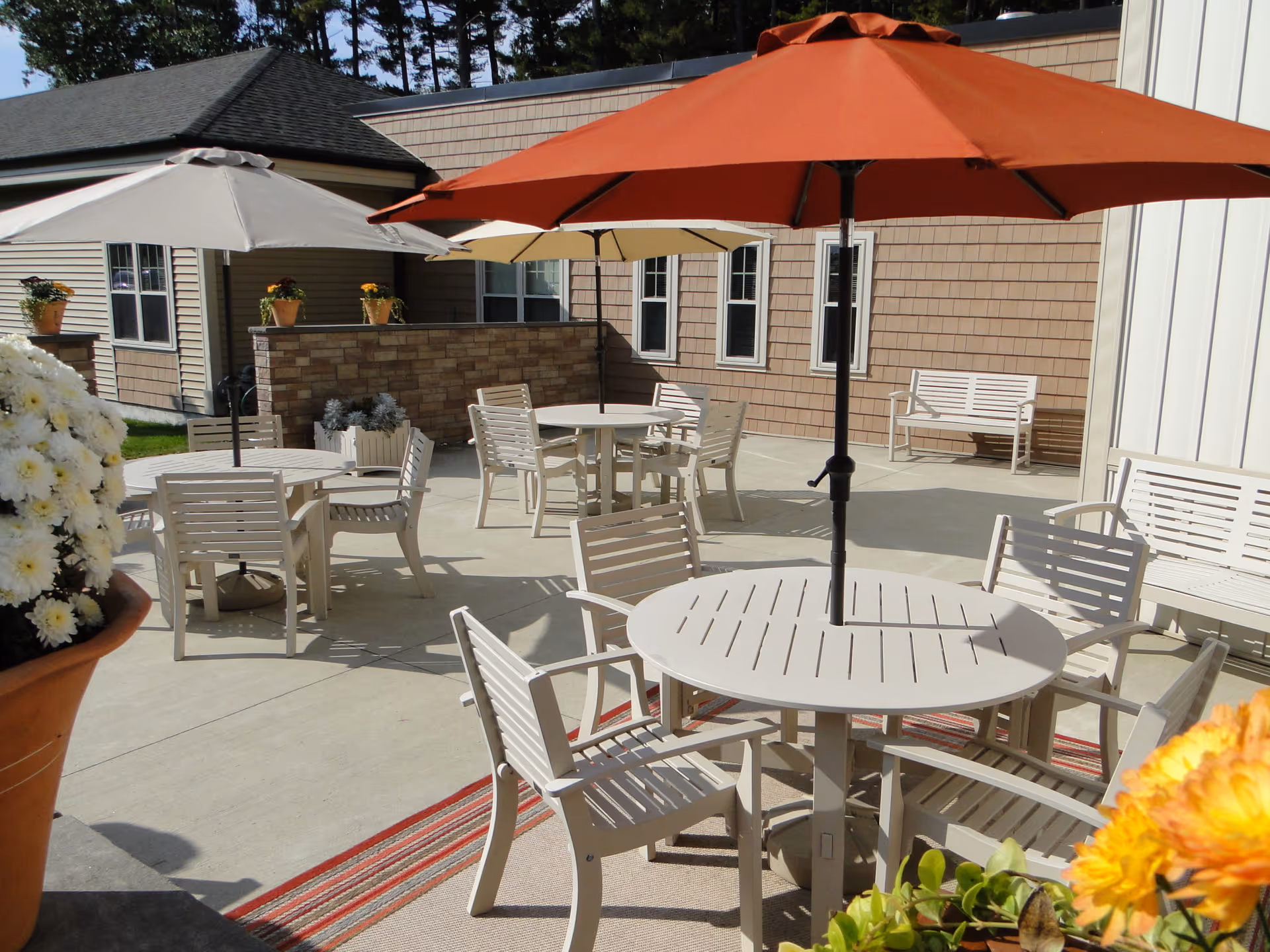 Outdoor patio area with several round tables and chairs, each table shaded by large umbrellas in beige, cream, and orange colors. There are potted flowers and plants around the patio, with a building featuring windows and siding in the background.