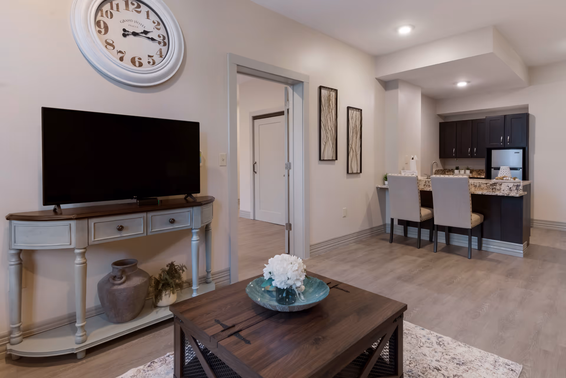 A modern living area featuring a flat-screen TV on a light-colored console table with decorative vases underneath. A large wall clock hangs above the TV. In the foreground, there is a wooden coffee table with a decorative bowl and white flowers. The room has light wood flooring and neutral-colored walls. To the right, there is a kitchen area with dark cabinets, a granite countertop, and two upholstered chairs at the counter. Two framed artworks hang on the wall near an open doorway leading to another room.