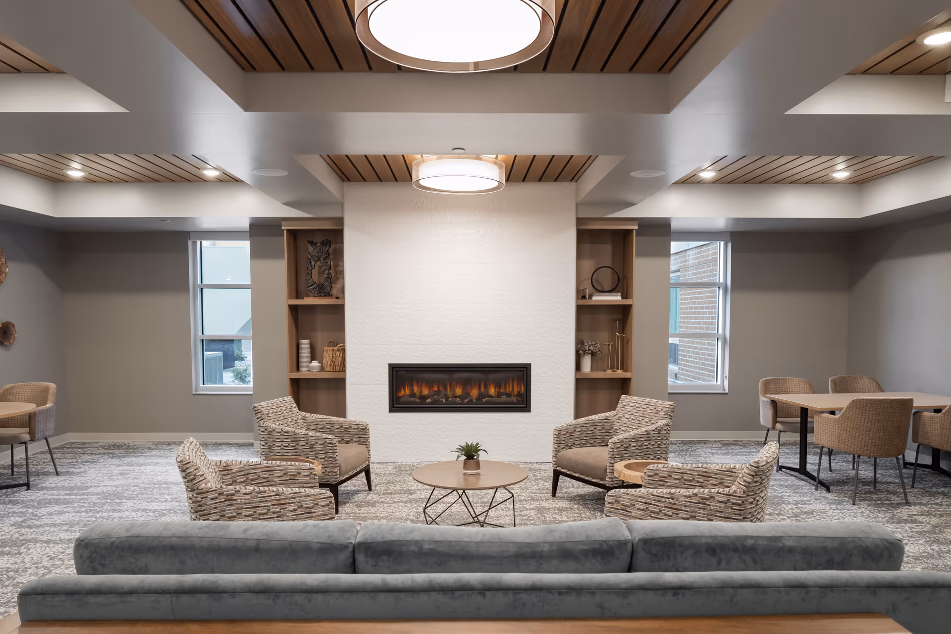 A modern lounge area with a gray sofa in the foreground, four patterned armchairs arranged around a round coffee table with a small plant, and a built-in electric fireplace set in a white textured wall flanked by wooden shelves. The ceiling features wood paneling and recessed lighting, and there are windows on either side of the fireplace letting in natural light. Additional seating with tables and chairs is visible on the sides.