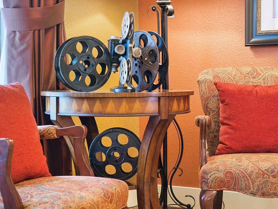 Two upholstered armchairs with red pillows flank a wooden side table displaying decorative vintage film reels against an orange wall.