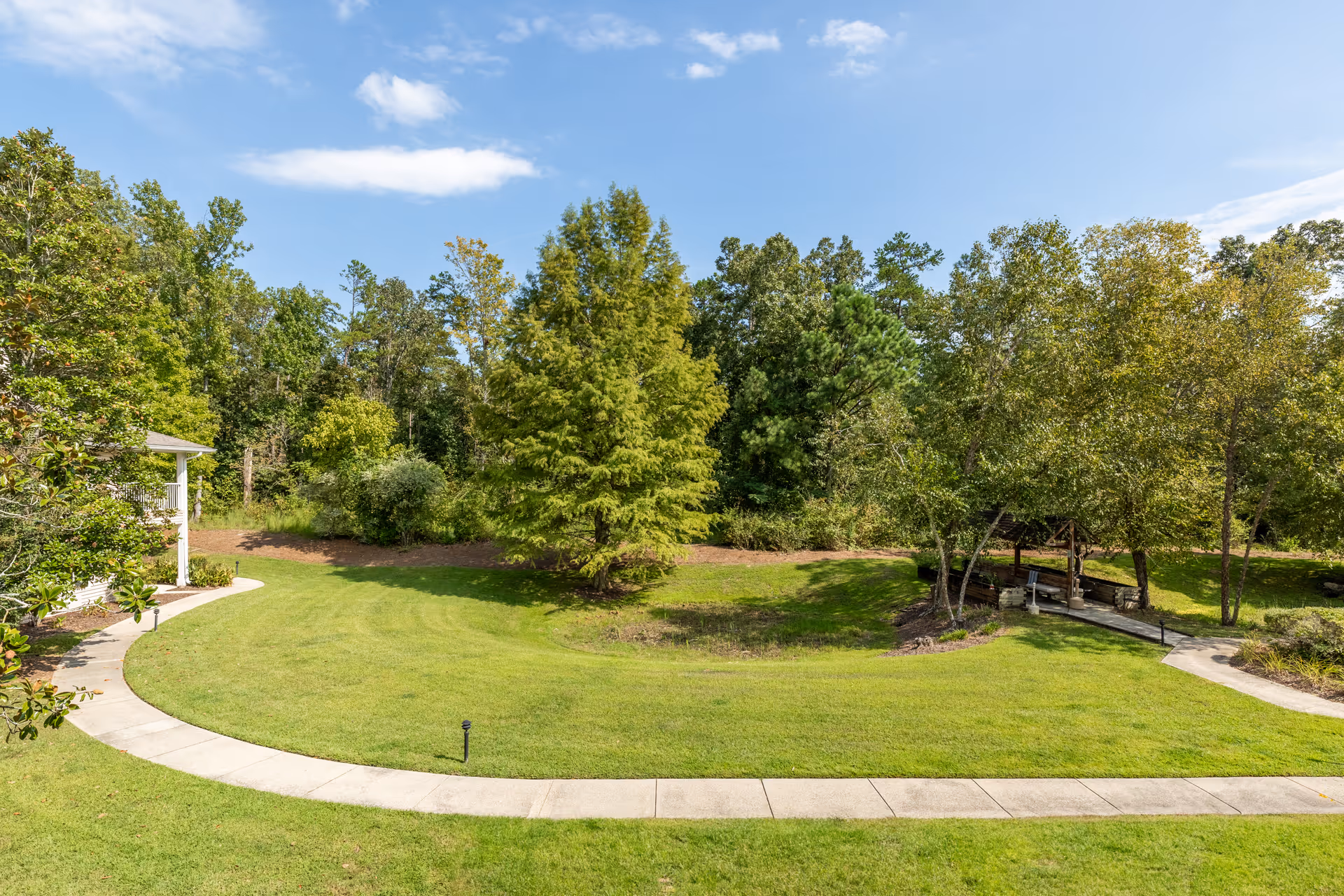 A well-maintained outdoor garden area with a curved concrete walkway, green grass, several trees, and a small wooden gazebo surrounded by trees under a partly cloudy blue sky.