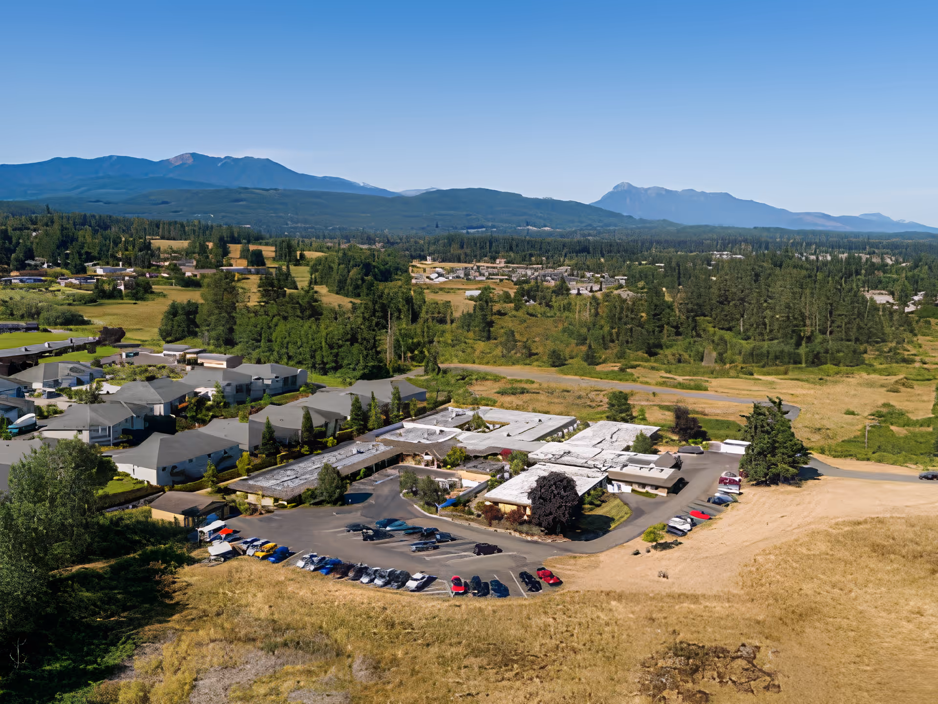 Aerial view of Avamere Olympic Rehabilitation of Sequim facility surrounded by greenery and open fields, with mountains visible in the background under a clear blue sky. The facility consists of multiple connected buildings with parking lots filled with cars.