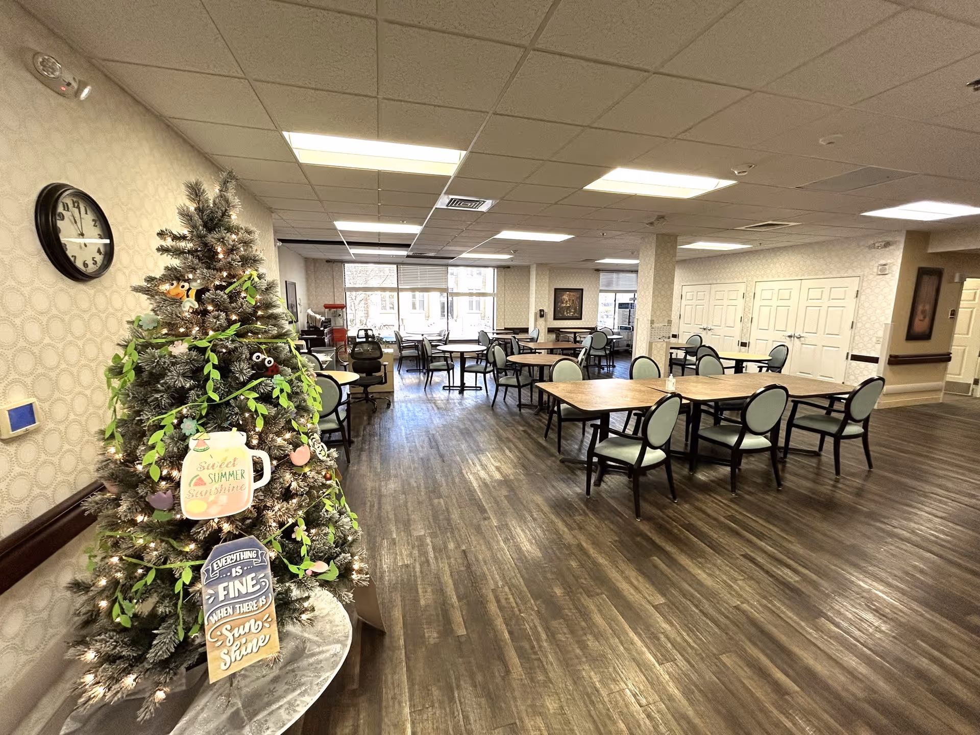 A spacious dining room in a senior living facility with multiple wooden tables and chairs arranged neatly. A decorated artificial tree with lights and summer-themed signs is placed on the left side near a wall clock. The room has large windows allowing natural light to brighten the space, and the floor is covered with wood-style flooring.
