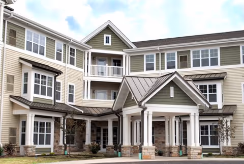 Exterior view of a multi-story residential building with beige and green siding, white trim, multiple windows, and a covered entrance supported by white columns.