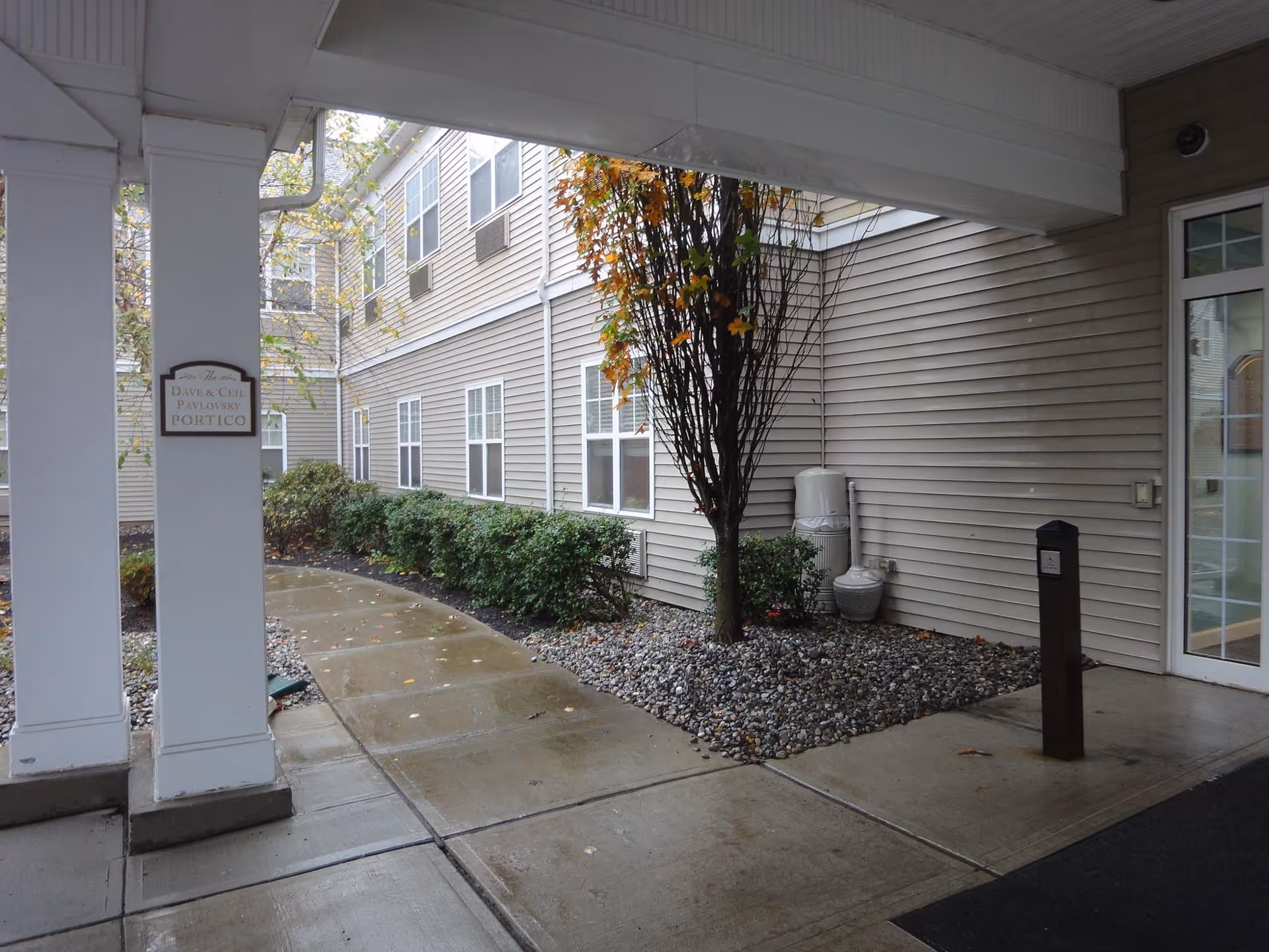 Covered walkway area outside a building with beige siding, a tree surrounded by rocks, bushes along the building, and a sign on a pillar reading 'The Dave & Chet Pavlovsky Portico'. The ground is wet, indicating recent rain.