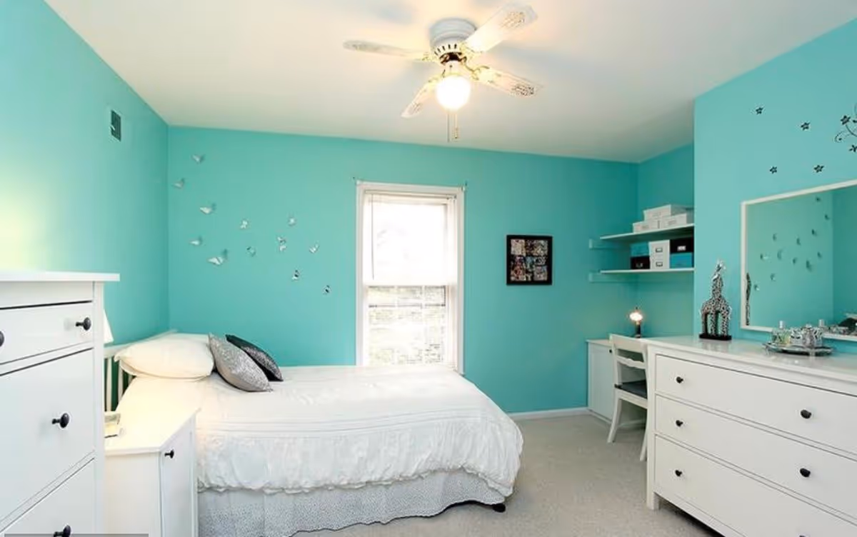 A bright bedroom with turquoise walls featuring a white bed with pillows, a white dresser, a white nightstand, a ceiling fan, a window with blinds, a desk with shelves holding boxes, and a large mirror on the wall.