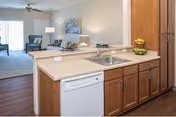 Open-concept kitchen with a sink, dishwasher, and wooden cabinets overlooking a living area with seating and sliding glass doors.