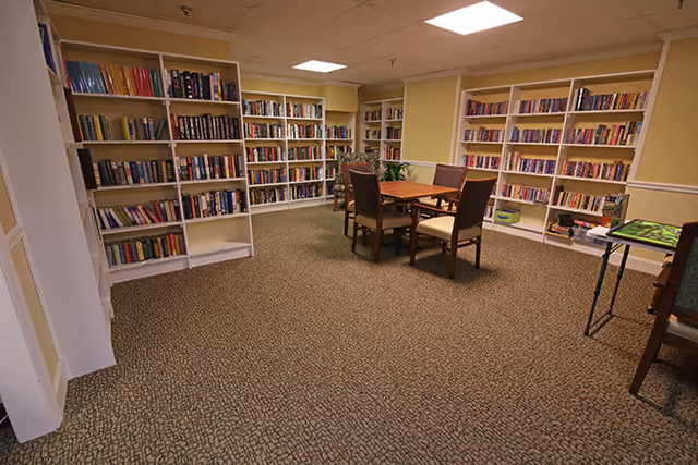 A cozy library room with multiple white bookshelves filled with books along the walls. In the center, there is a wooden table with four chairs around it. The room has beige walls and a patterned carpeted floor, with ceiling lights providing illumination.