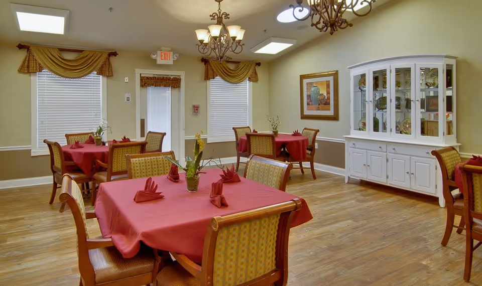 A dining room with wooden floors and several tables covered with red tablecloths and matching folded napkins. Each table has four chairs with patterned upholstery. The room features two windows with blinds and gold valances, a white china cabinet displaying decorative items, a framed painting on the wall, and two ornate chandeliers hanging from the ceiling.