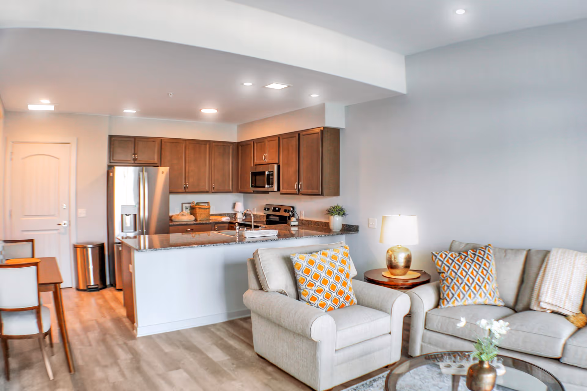 Open-plan living area with a sofa and armchair by a coffee table and a kitchen with an island and stainless steel appliances in the background.