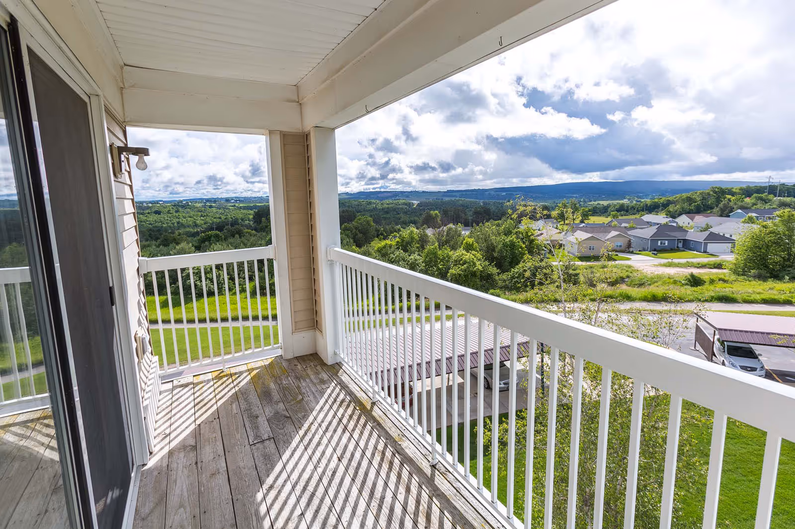 View from a covered balcony with white railings and wooden floorboards, overlooking a green landscape with trees, houses, and distant hills under a partly cloudy sky.