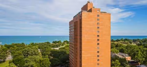 Tall brick senior living building rises beside trees with a blue ocean backdrop.