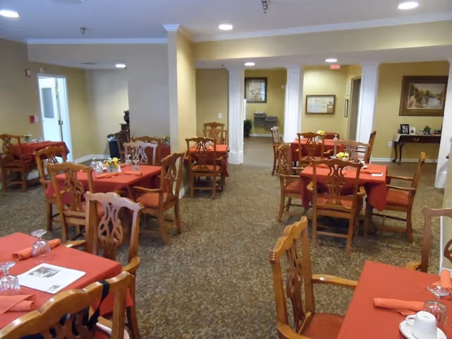 Interior view of a dining room with multiple wooden tables and chairs. The tables are covered with red tablecloths and set with glasses, napkins, and small flower arrangements. The room has beige walls, carpeted floors, and recessed lighting in the ceiling. There are doorways leading to other rooms in the background.