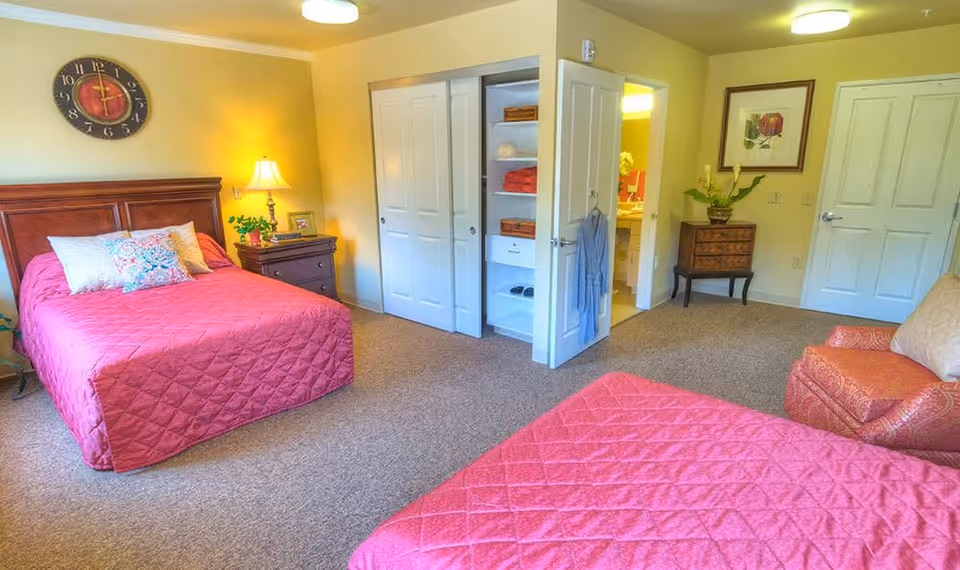 A bright bedroom with two beds covered in matching red quilted bedspreads. There is a wooden nightstand with a lamp and a small plant between the beds. A large wall clock hangs above one bed. The room has beige carpet and light yellow walls. An open closet with shelves holding towels and baskets is visible, as well as a doorway leading to a bathroom. A framed floral picture hangs on the wall near a small wooden chest and a red patterned armchair with a beige pillow.