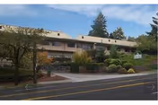 Exterior view of Park Rose Care Center, a two-story building with a beige facade surrounded by trees and shrubs, situated along a road with a sidewalk.