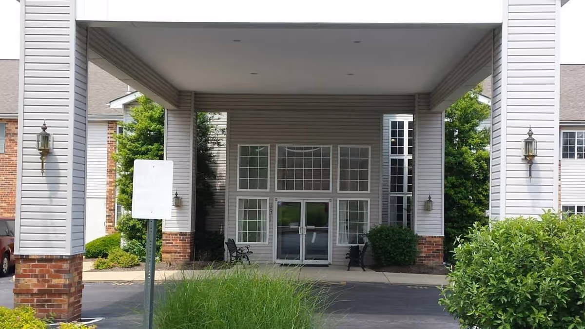 Entrance of a senior living facility with a covered drop-off area supported by large white columns with brick bases. The building has large windows and double glass doors. There are benches and greenery around the entrance.