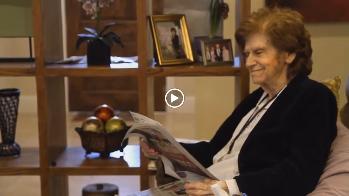 An elderly woman sitting comfortably on a couch in a cozy living room area, reading a newspaper. Behind her is a wooden shelving unit with decorative items including framed photos, a plant, and ornamental balls.
