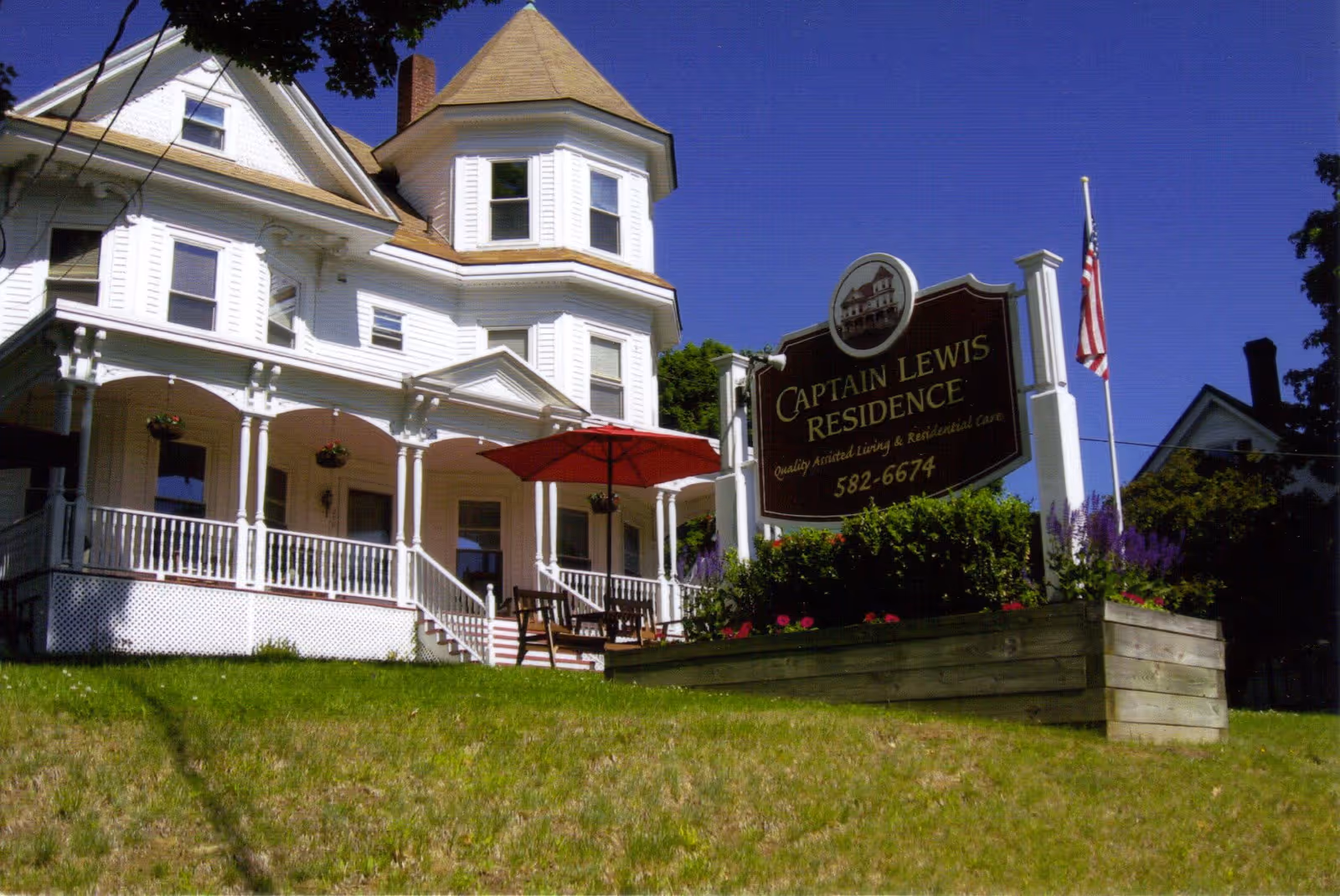 Victorian-style white residence with a wraparound porch, a red patio umbrella, and a sign reading "Captain Lewis Residence" on a grassy lawn.