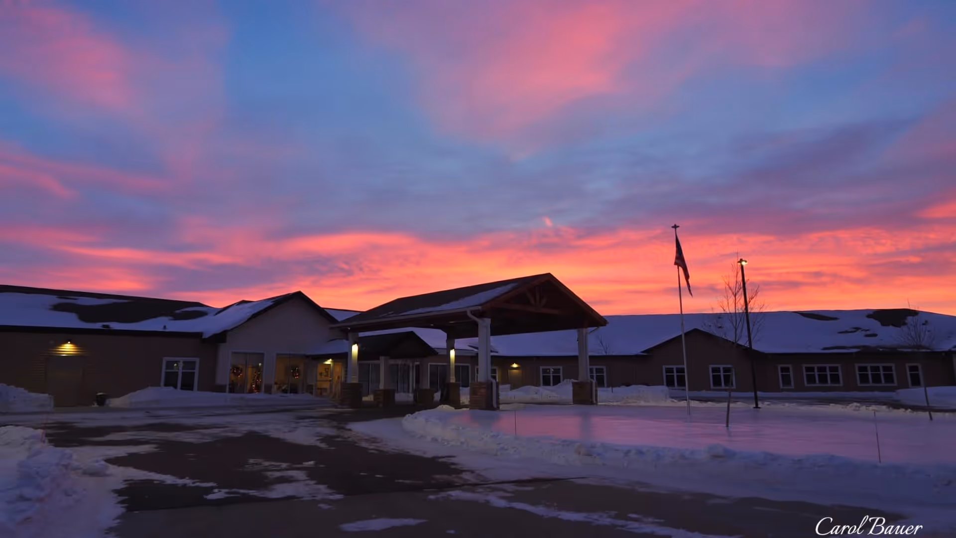 Front entrance of a senior living building under a colorful sunset sky with snow on the ground and an American flag.