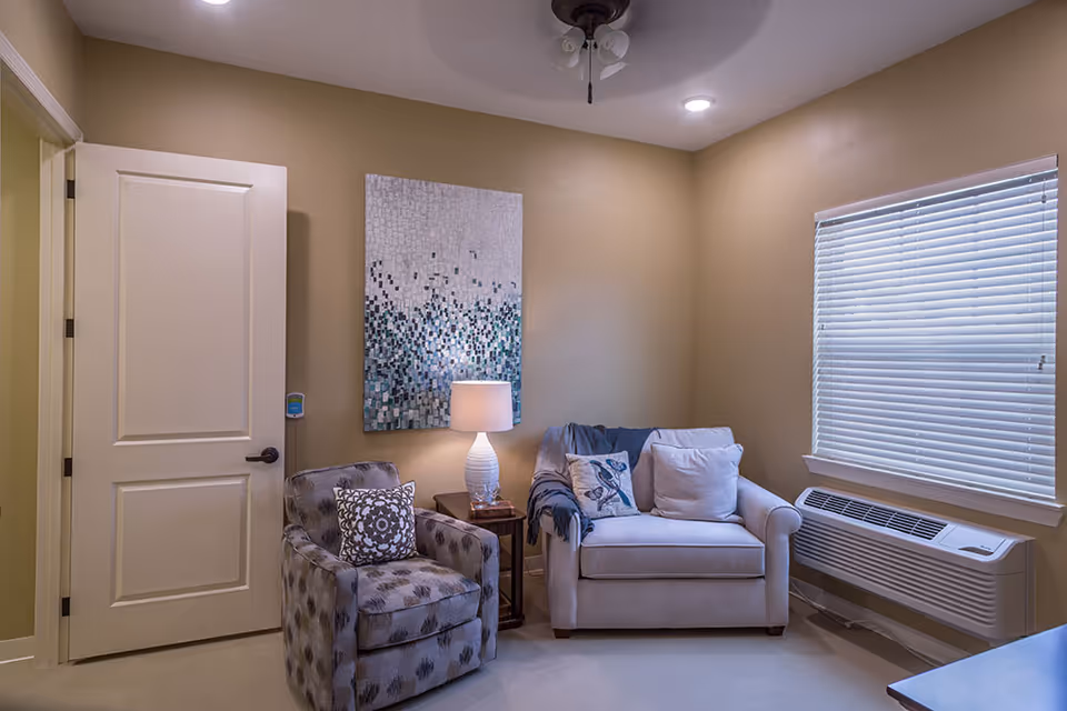 A cozy living room with beige walls featuring a patterned armchair and a light-colored loveseat with decorative pillows. A small wooden side table with a white lamp sits between the chairs. A large window with closed blinds is on the right wall, and a ceiling fan with lights is visible above. A piece of abstract artwork hangs on the wall behind the side table.