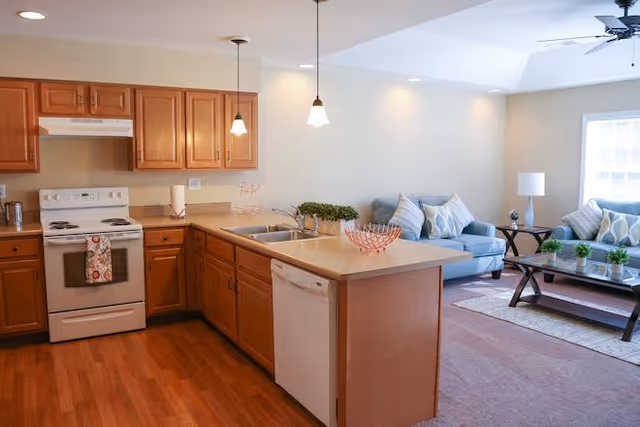 A bright and clean open kitchen and living room area. The kitchen features wooden cabinets, a white stove with a towel hanging on the handle, a dishwasher, and a sink on the island countertop. The living room has two blue sofas with patterned cushions, a glass coffee table, a side table with a lamp, and a ceiling fan. The space is well-lit with recessed lighting and natural light from a window.