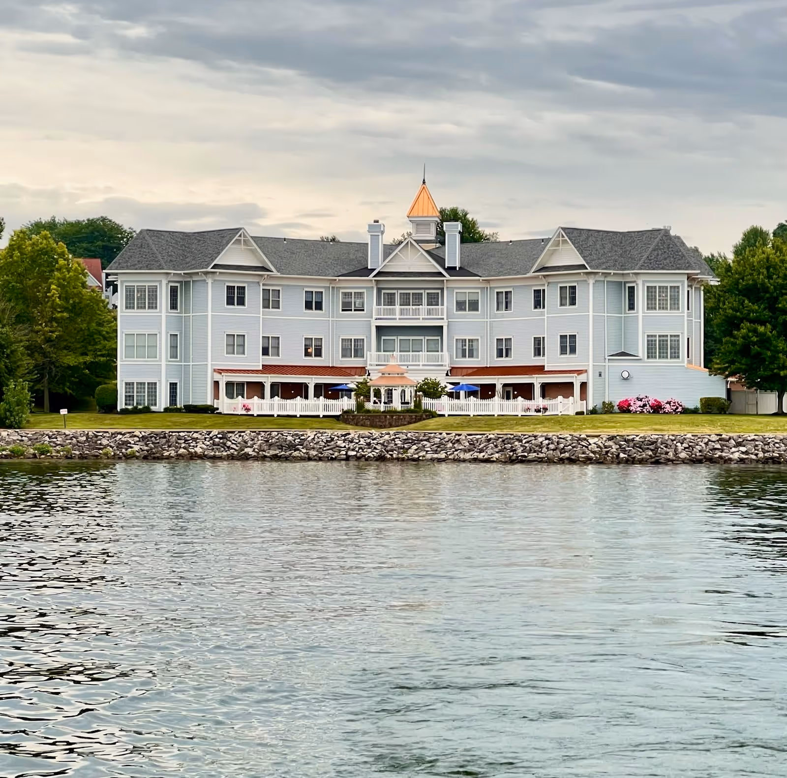 A large, light blue multi-story building with a peaked roof and a small cupola on top, situated beside a body of water with a rocky shoreline. The building has multiple windows, a white picket fence, a small gazebo, and some outdoor seating with blue umbrellas. Trees and greenery surround the building under a cloudy sky.