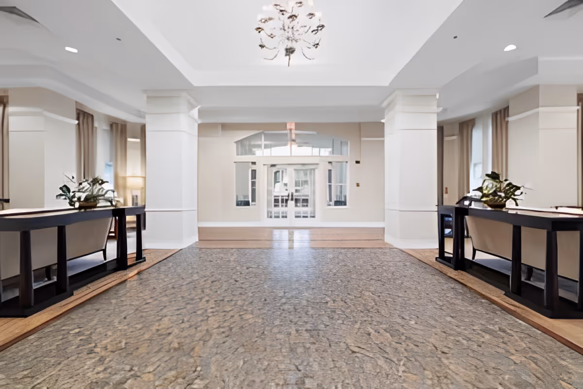 Spacious and well-lit lobby area with a textured stone floor, white walls, large columns, and two dark wooden tables with plants on either side. A chandelier hangs from the ceiling, and large windows and glass doors are visible at the far end.