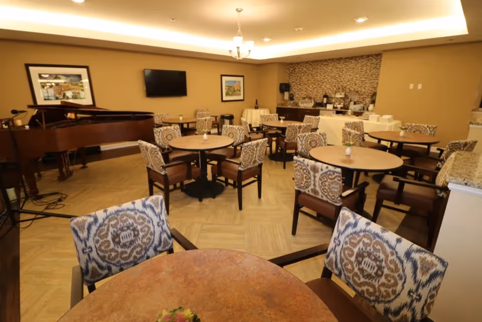 Dining/activity room with round tables and patterned chairs, a grand piano, wall-mounted TV, and a buffet counter.
