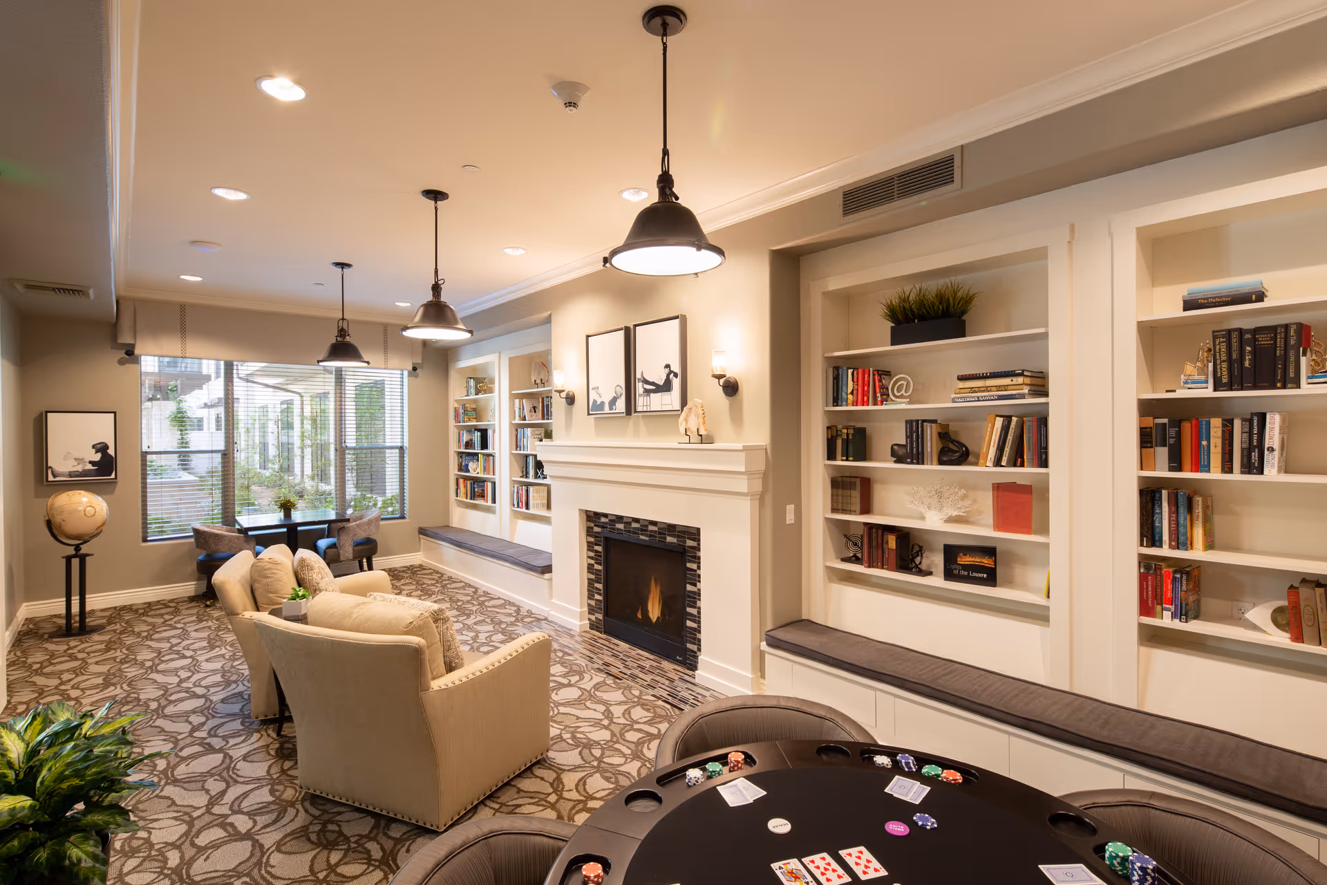 Cozy lounge with armchairs, a fireplace flanked by built-in bookshelves, pendant lights, large window seating area, and a poker table in the foreground.