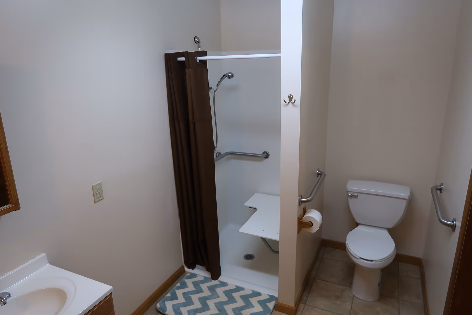 A bathroom with a white toilet, a shower area with a brown curtain and a foldable white shower seat, grab bars on the walls, a sink with a wooden cabinet, and a blue and white chevron patterned bath mat on the floor.