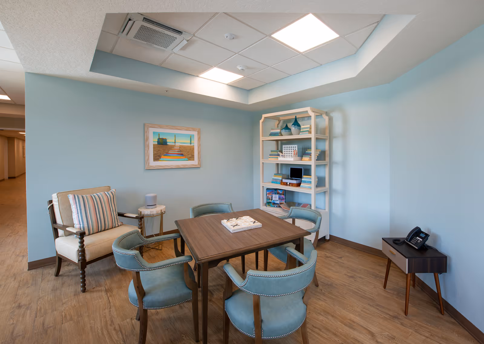 A cozy sitting area in a senior living facility with a wooden table surrounded by four teal upholstered chairs. There is a beige armchair with a striped cushion next to a small round side table. A white bookshelf filled with books and decorative items is against the light blue wall. A framed picture of a beach scene hangs on the wall above the armchair. The room has wood flooring and a ceiling with recessed lighting.