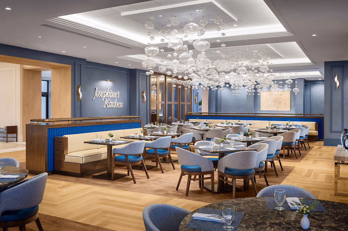 A spacious and elegant dining room with multiple round tables set with glassware, napkins, and small floral centerpieces. The room features light wood flooring, blue and beige upholstered chairs, and a long beige banquette with blue paneling. A modern chandelier with numerous hanging glass orbs is suspended from the ceiling. The wall has a sign that reads 'Josephine's Kitchen'.