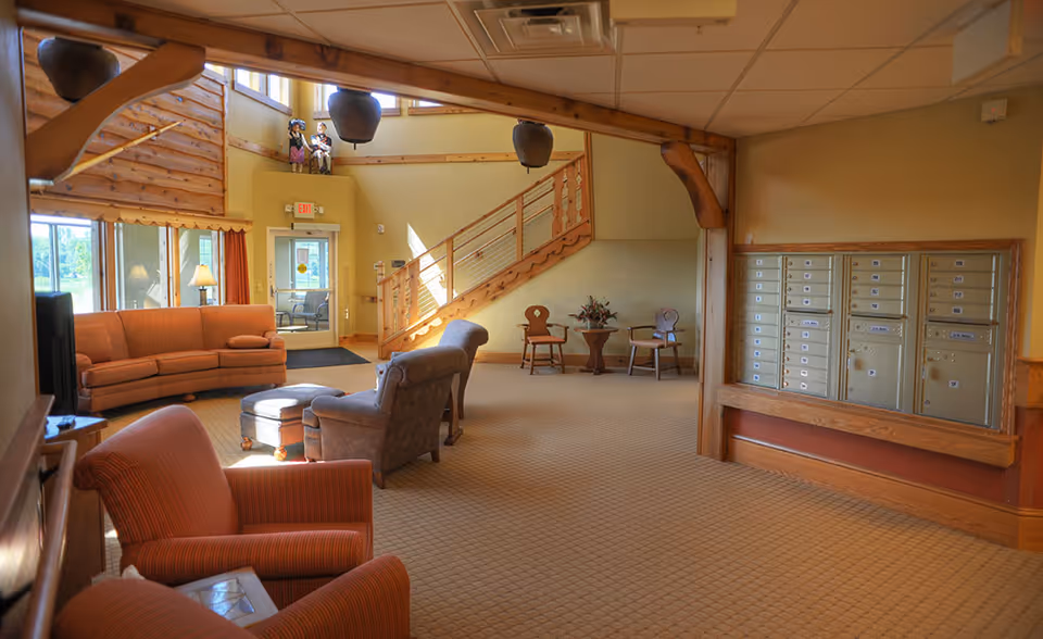 A sunlit assisted-living lobby with sofas and armchairs, a wooden staircase, and a wall of resident mailboxes.