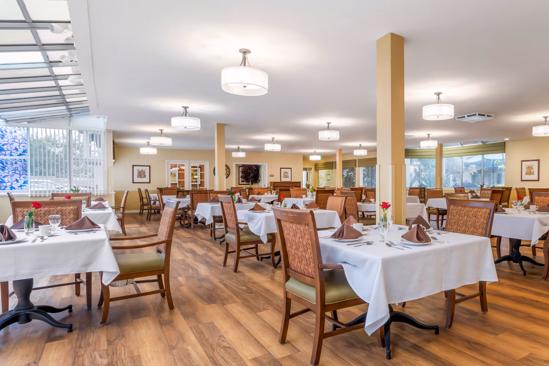 A spacious dining room with multiple tables covered in white tablecloths, each set with brown napkins, glassware, and silverware. The room features wooden chairs with patterned upholstery, wood flooring, and several ceiling lights. Large windows and a glass ceiling section allow natural light to fill the space.