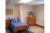 A simple, clean bedroom with a single wooden bed covered with a blue bedspread and a striped pillow. There is a wooden nightstand next to the bed and a wooden dresser with a vase of flowers on top. The room has beige walls, a bulletin board, and a curtain that can be drawn for privacy. The floor is light-colored with a dark rug near the bed.