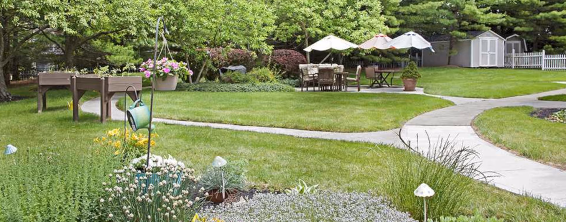 A well-maintained outdoor garden area with green grass, flowering plants, and shrubs. There are paved walkways curving through the garden, leading to a seating area with tables, chairs, and umbrellas. In the background, there are trees, a white picket fence, and a small shed.
