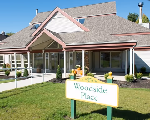 Exterior view of a senior living facility building with a peaked roof and large windows. In front of the building is a green lawn with a sign that reads 'Woodside Place'. There are some small shrubs and plants near the building entrance.