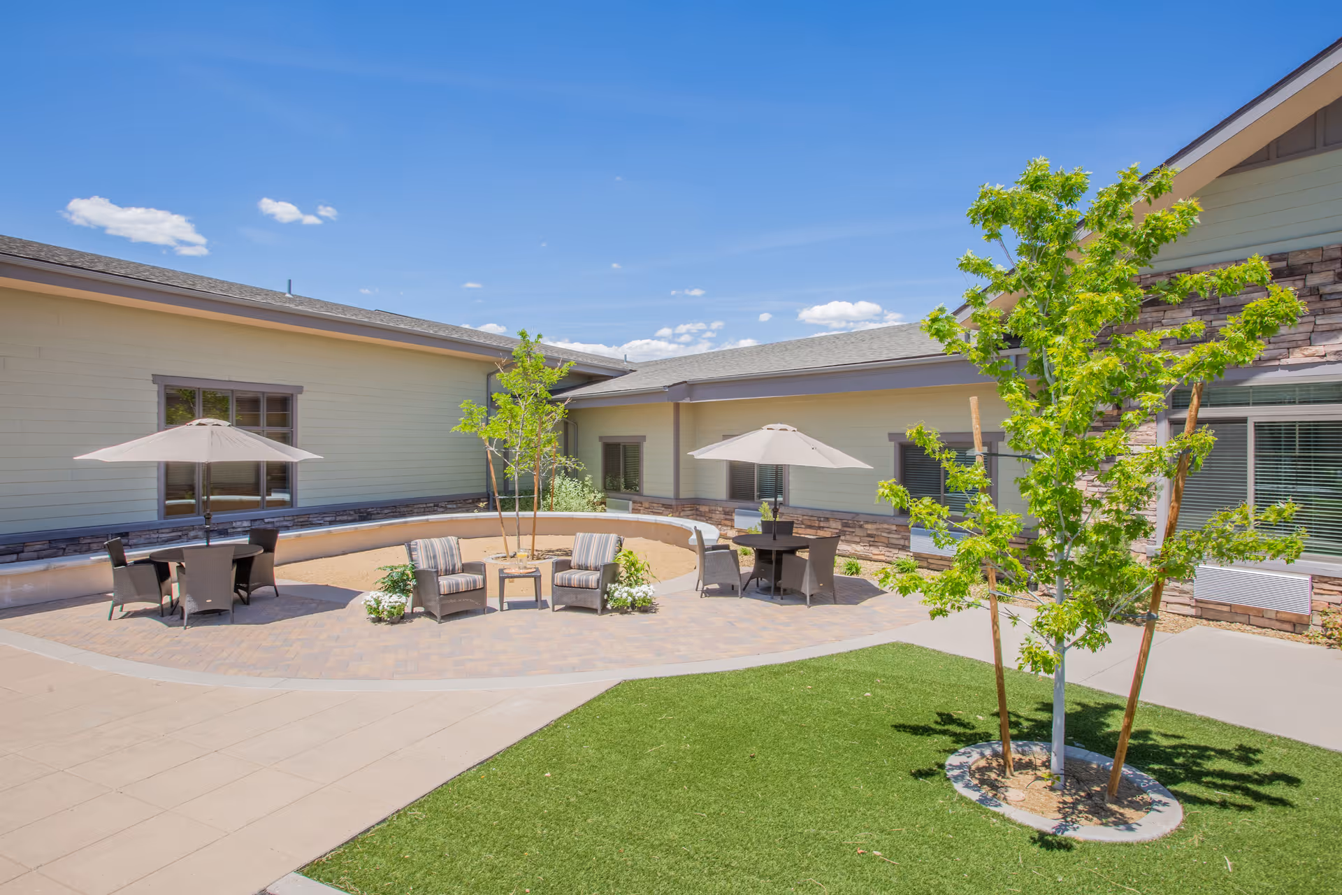 Outdoor courtyard area at Aspire Transitional Care featuring patio tables with umbrellas, cushioned chairs, small trees, and a well-maintained lawn under a clear blue sky.