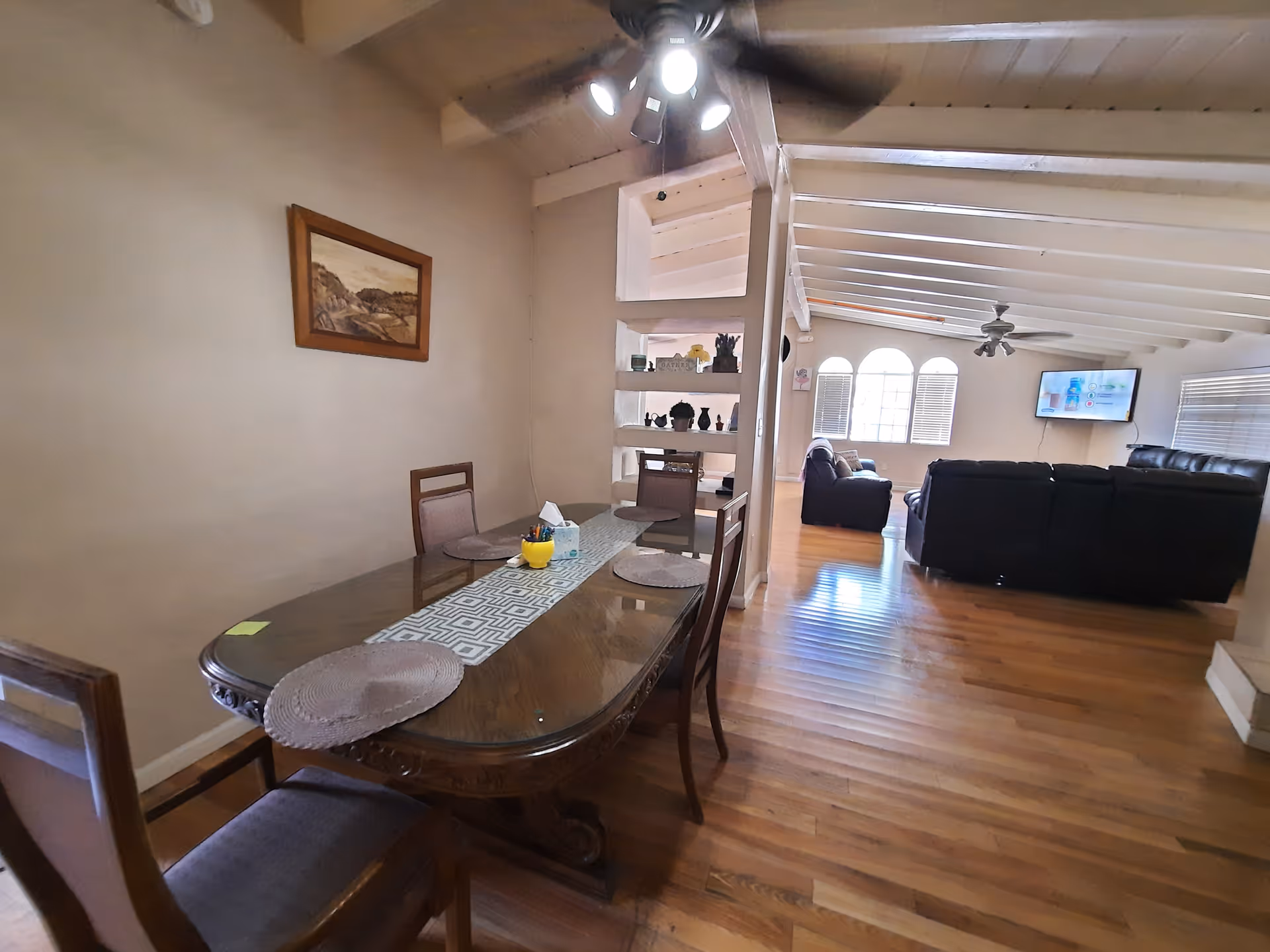 Dining area with a wooden table and chairs that opens into a living room with sofas and a wall-mounted TV under a beamed ceiling.