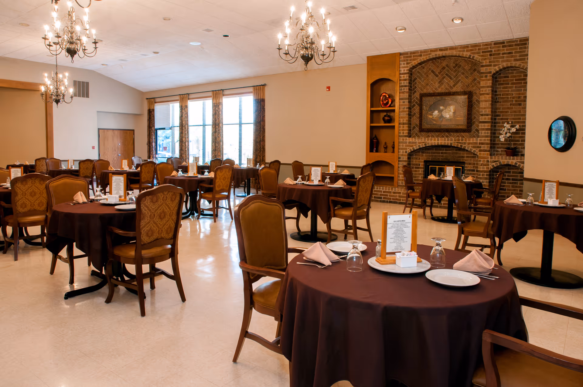 A dining room with multiple round tables covered in dark brown tablecloths, each set with plates, glasses, napkins, and menus. The room features upholstered chairs, chandeliers hanging from the ceiling, large windows with curtains, and a brick fireplace with decorative shelves.