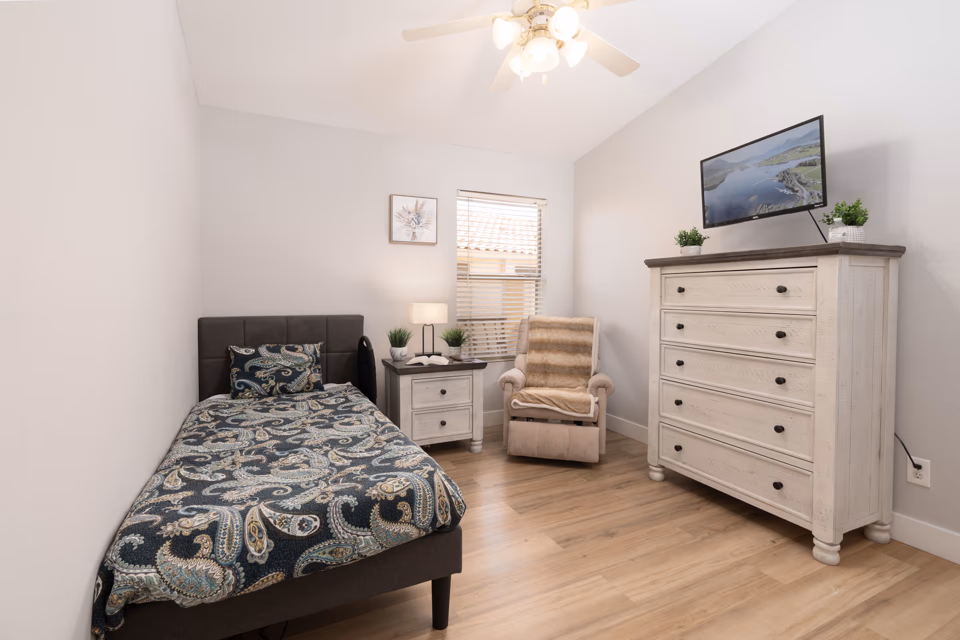 A small, neatly arranged bedroom with a single bed covered in a patterned blue and beige bedspread, a nightstand with a lamp and small plants, a beige recliner chair with a throw blanket, a window with blinds, and a white wooden dresser with a flat-screen TV mounted above it.