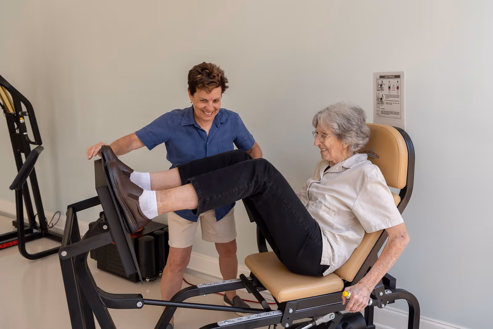 An elderly woman is exercising on a leg press machine with the assistance of a smiling caregiver in a fitness room. The elderly woman is seated and pushing against the footplate, while the caregiver stands nearby offering support.
