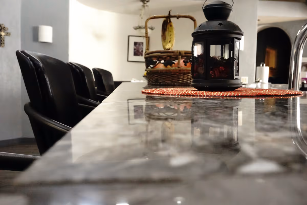Close-up view of a polished granite countertop with a black lantern and a woven basket holding fruit on top. In the background, there are black cushioned chairs lined up along the counter, a wall with a framed picture, and a cross decoration.