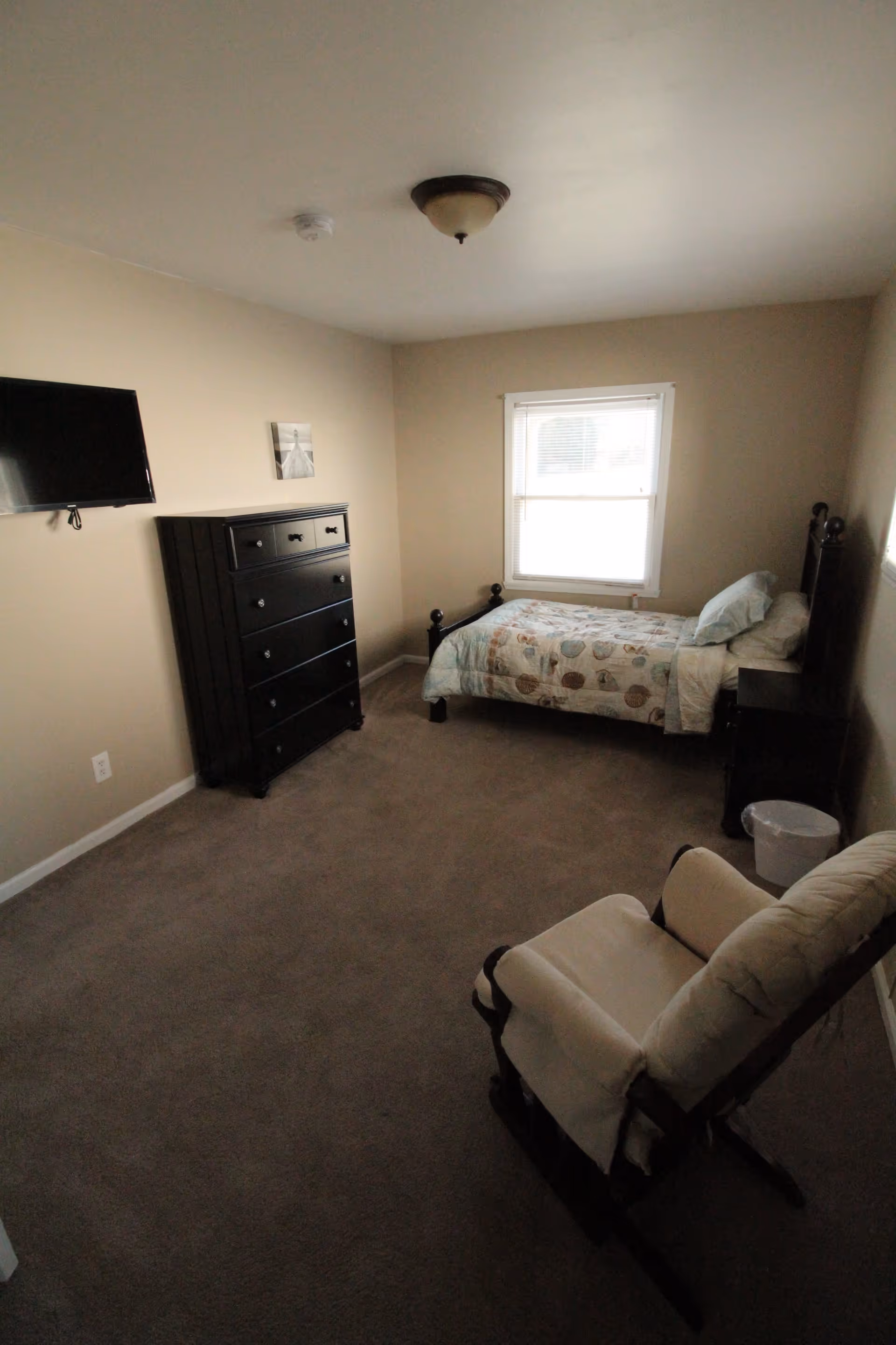 A small bedroom with beige walls and carpet. The room contains a single bed with a patterned comforter, a black dresser, a wall-mounted flat-screen TV, a cushioned rocking chair, a small nightstand, and a window with blinds letting in natural light.