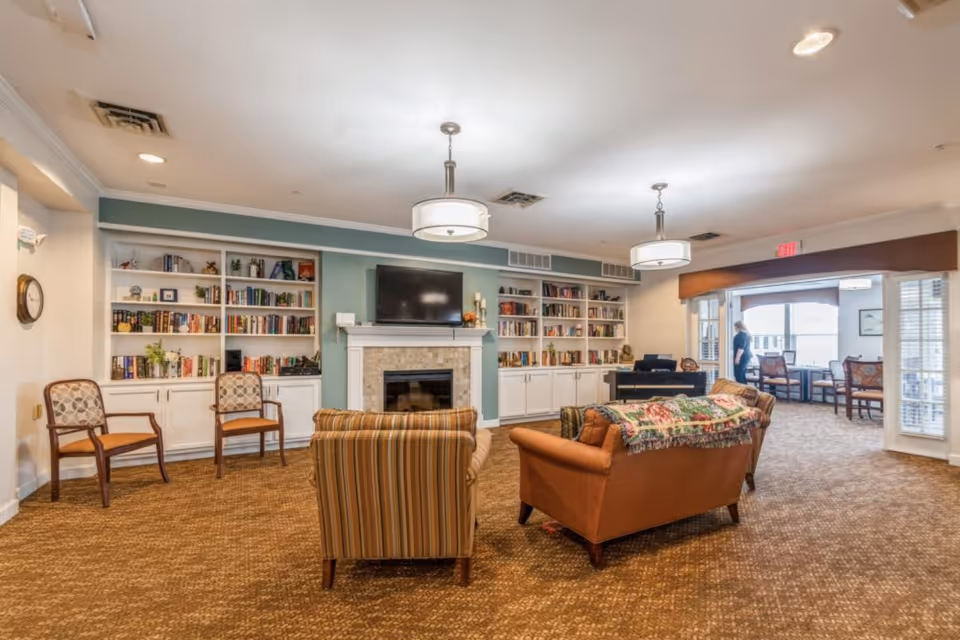A cozy living room area in a senior living facility with a fireplace and a mounted TV above it. The room features built-in white bookshelves filled with books and decorative items on either side of the fireplace. There are two armchairs and a sofa with a colorful throw blanket arranged around the fireplace. The carpet is brown, and the walls are painted in soft green and white tones. Two ceiling lights hang from the ceiling, and a person is visible in the background near a windowed seating area.