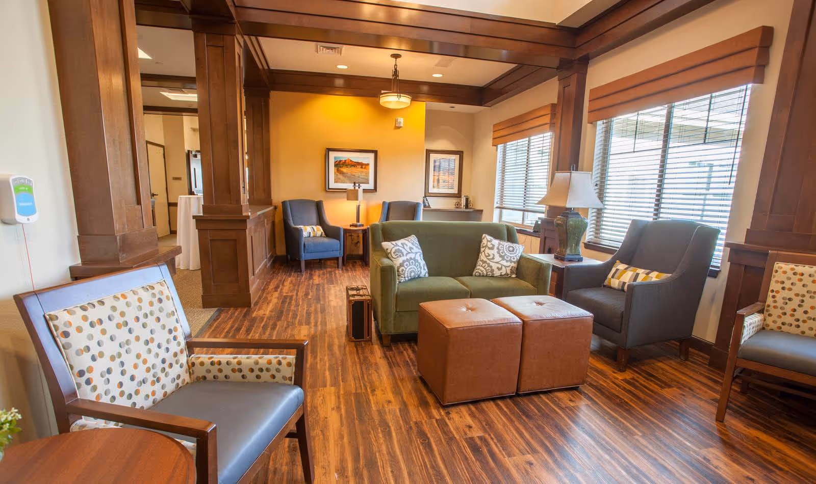 A cozy living room area in a senior living facility with wooden flooring and wood-paneled columns. The room features a green loveseat with two patterned pillows, two brown ottomans, a gray armchair with a striped pillow, and two blue armchairs with a small table and lamp between them. Large windows with wooden blinds allow natural light to fill the space. The walls are decorated with framed artwork and a warm yellow accent wall.