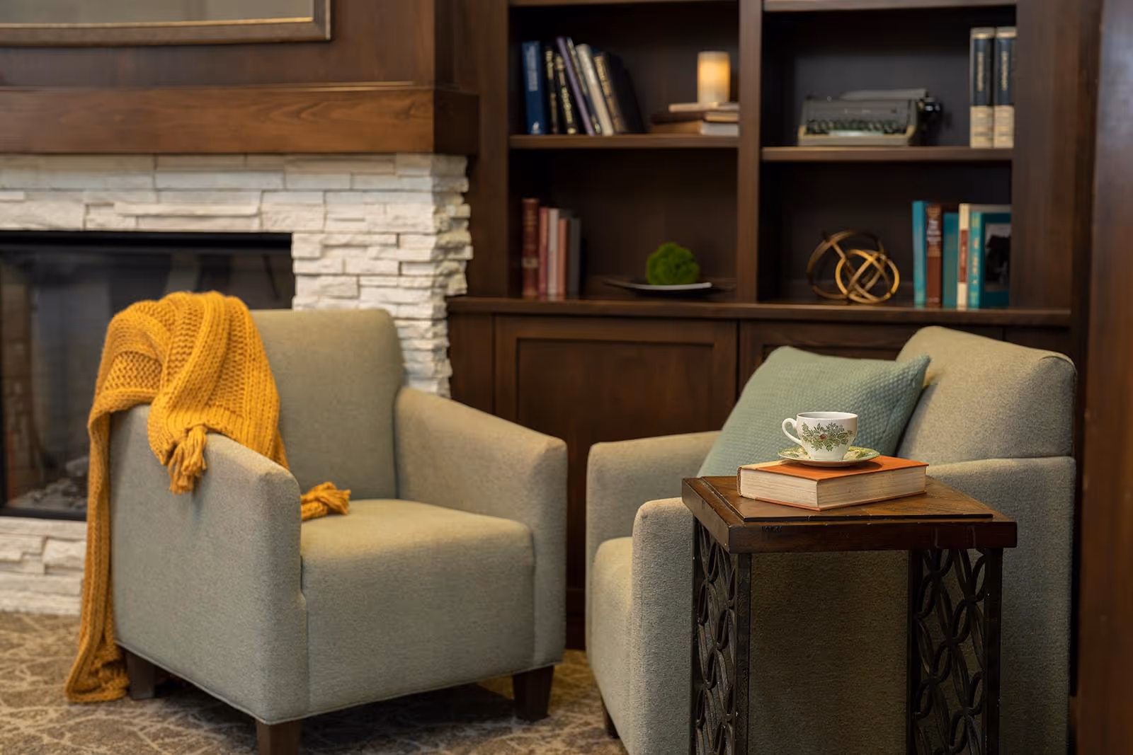 Two comfortable armchairs in a cozy living room setting with a mustard yellow knitted throw draped over one chair. A small wooden side table between the chairs holds a book and a teacup with a saucer. Behind the chairs is a built-in dark wood bookshelf filled with books and decorative items, and to the left is a stone fireplace.