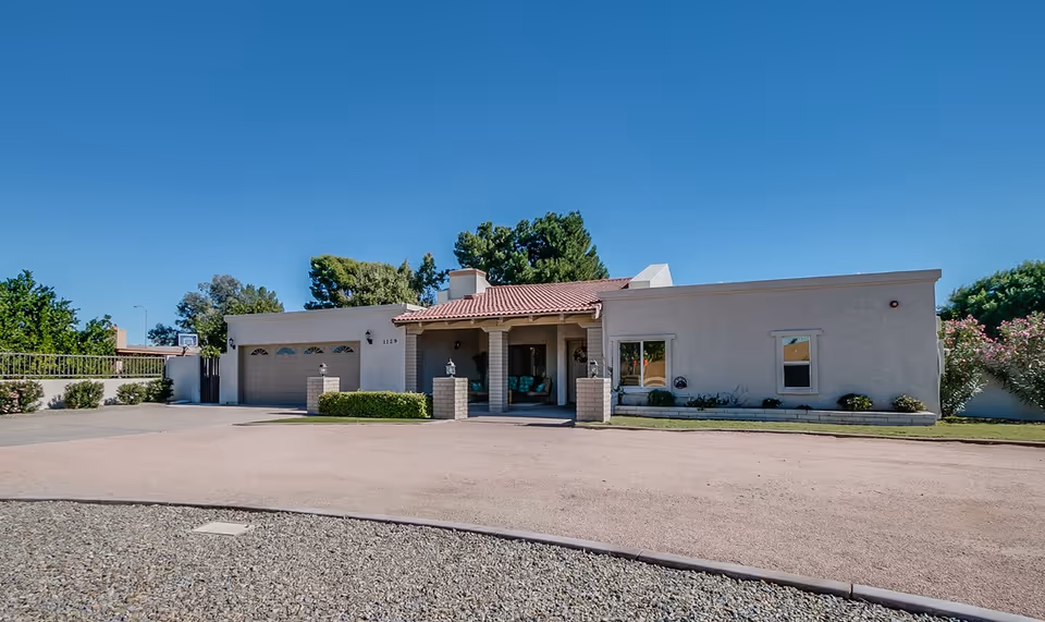 Single-story building with a beige exterior and a red tile roof under a clear blue sky. The building has a covered entrance with two brick pillars and a seating area visible inside. There is a two-car garage on the left side and a gravel driveway in front. Trees and shrubs surround the property.