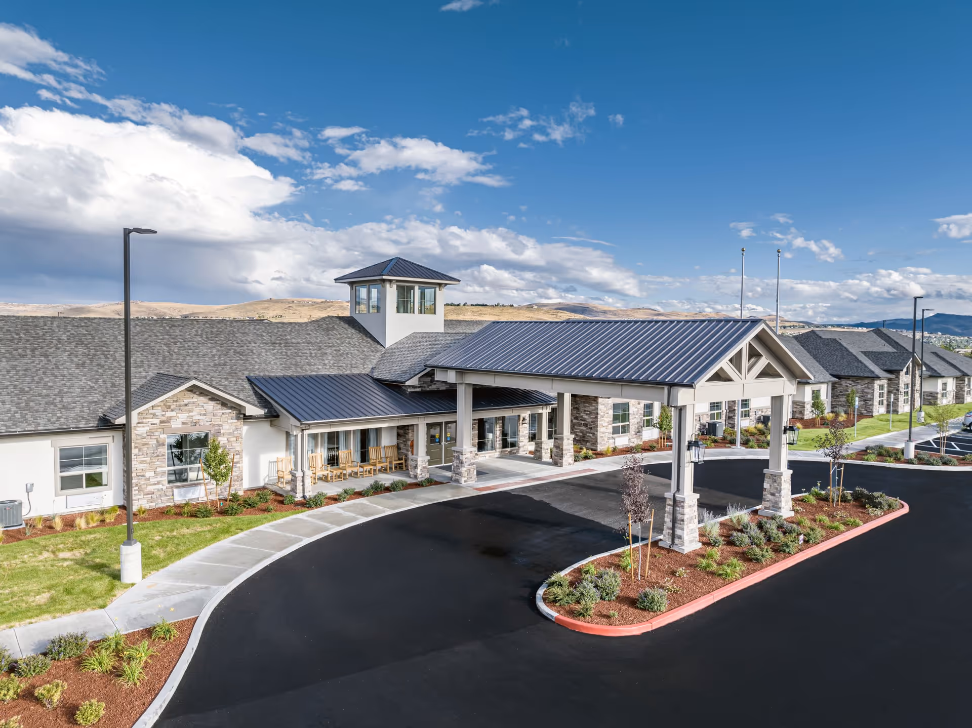 Exterior view of Kiley Ranch Senior Living facility showing a large building with stone and white walls, a covered entrance with a metal roof, several rocking chairs on the porch, landscaped greenery, and a clear blue sky with some clouds.
