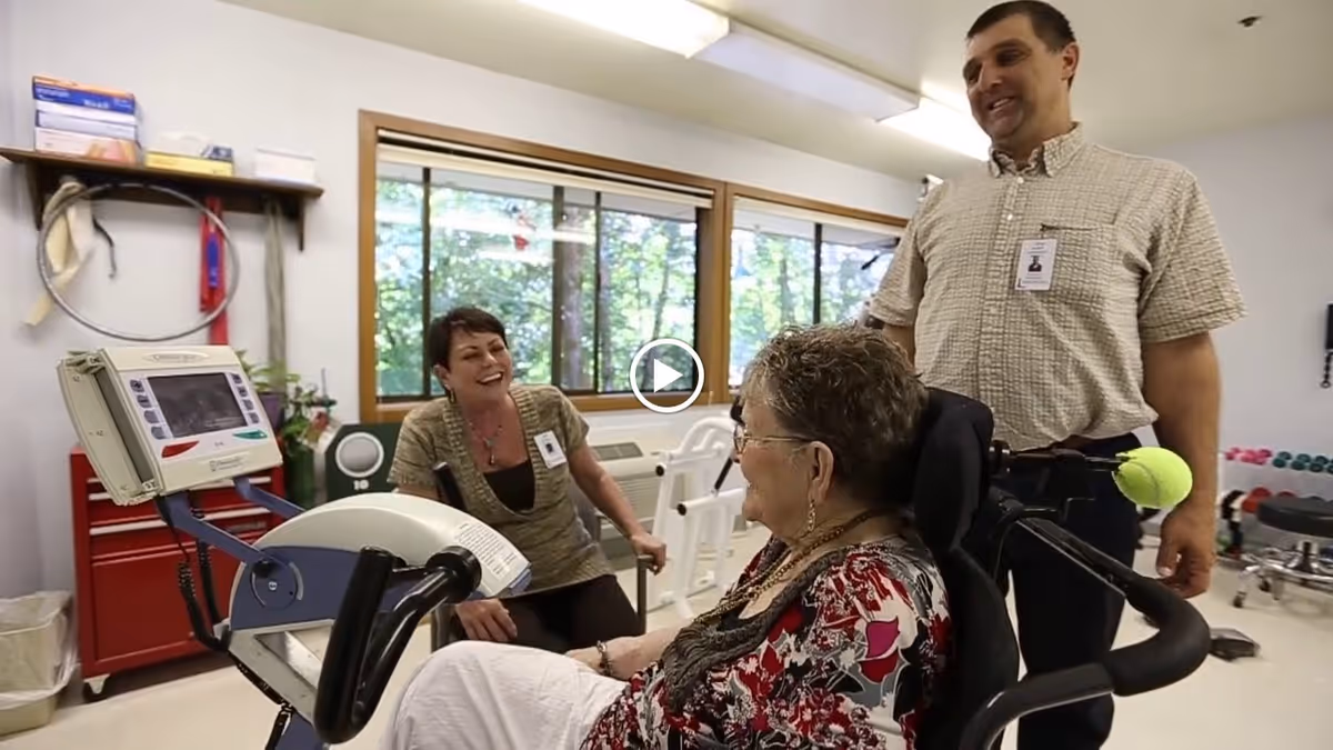 An elderly woman in a wheelchair is interacting with two staff members in a rehabilitation therapy room. The room has exercise equipment, large windows showing greenery outside, and a bright, clean environment.
